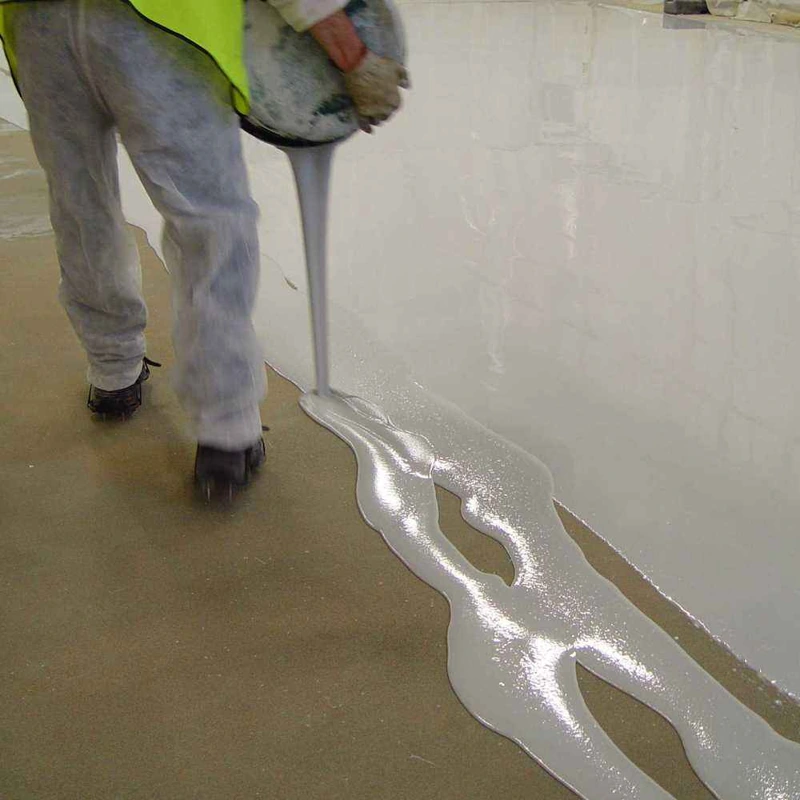 worker pouring light grey self-leveling epoxy resin onto a floor surface.