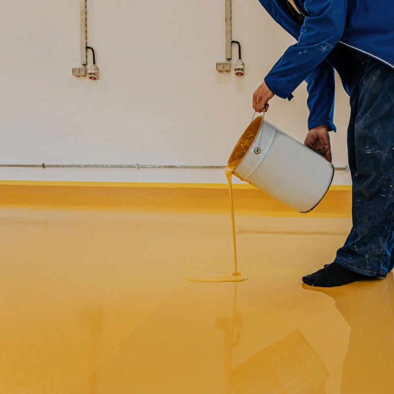worker pouring vibrant yellow self-leveling epoxy resin onto an indoor floor.